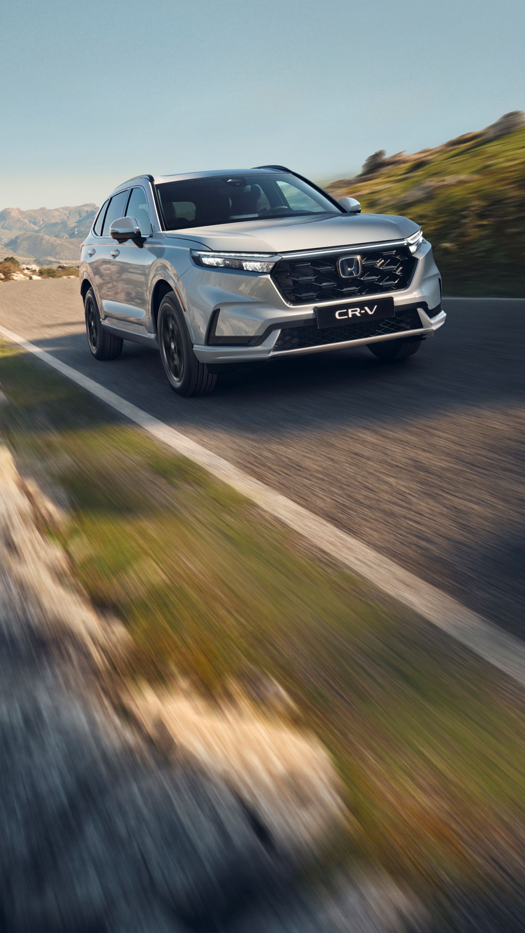 Un SUV Honda CR-V plateado conduciendo por una carretera de montaña sinuosa con terreno rocoso y montañas de fondo. El vehículo está en movimiento, creando una sensación de velocidad y aventura en el paisaje escarpado.