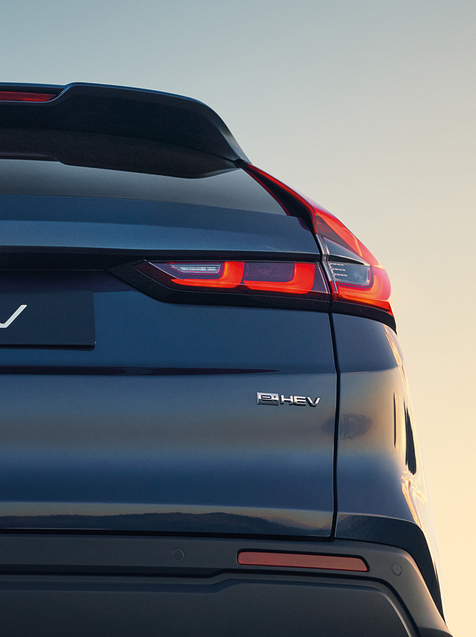 Primer plano trasero de un Honda CR-V e:HEV azul oscuro mostrando el emblema Honda, las letras CR-V y los pilotos traseros LED distintivos e iluminados, con el suave cielo del atardecer de fondo.