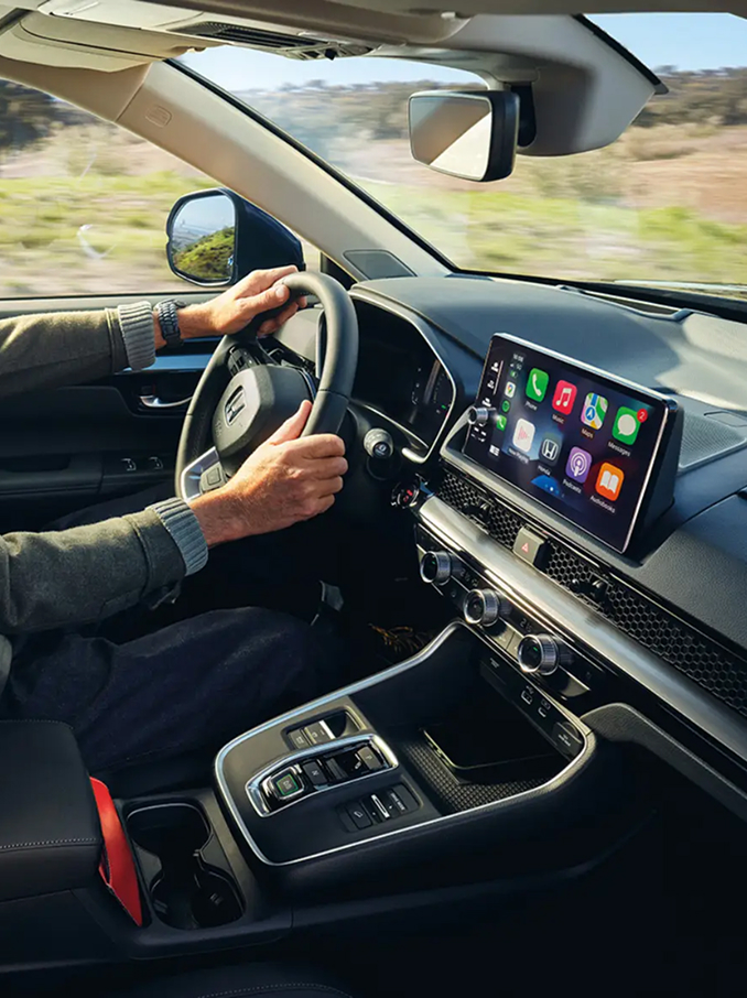 Vista interior de un coche moderno con un conductor sentado al volante, mostrando el salpicadero, el volante, la consola central y una amplia pantalla táctil con coloridos iconos de aplicaciones, mientras el paisaje rural pasa por las ventanillas.