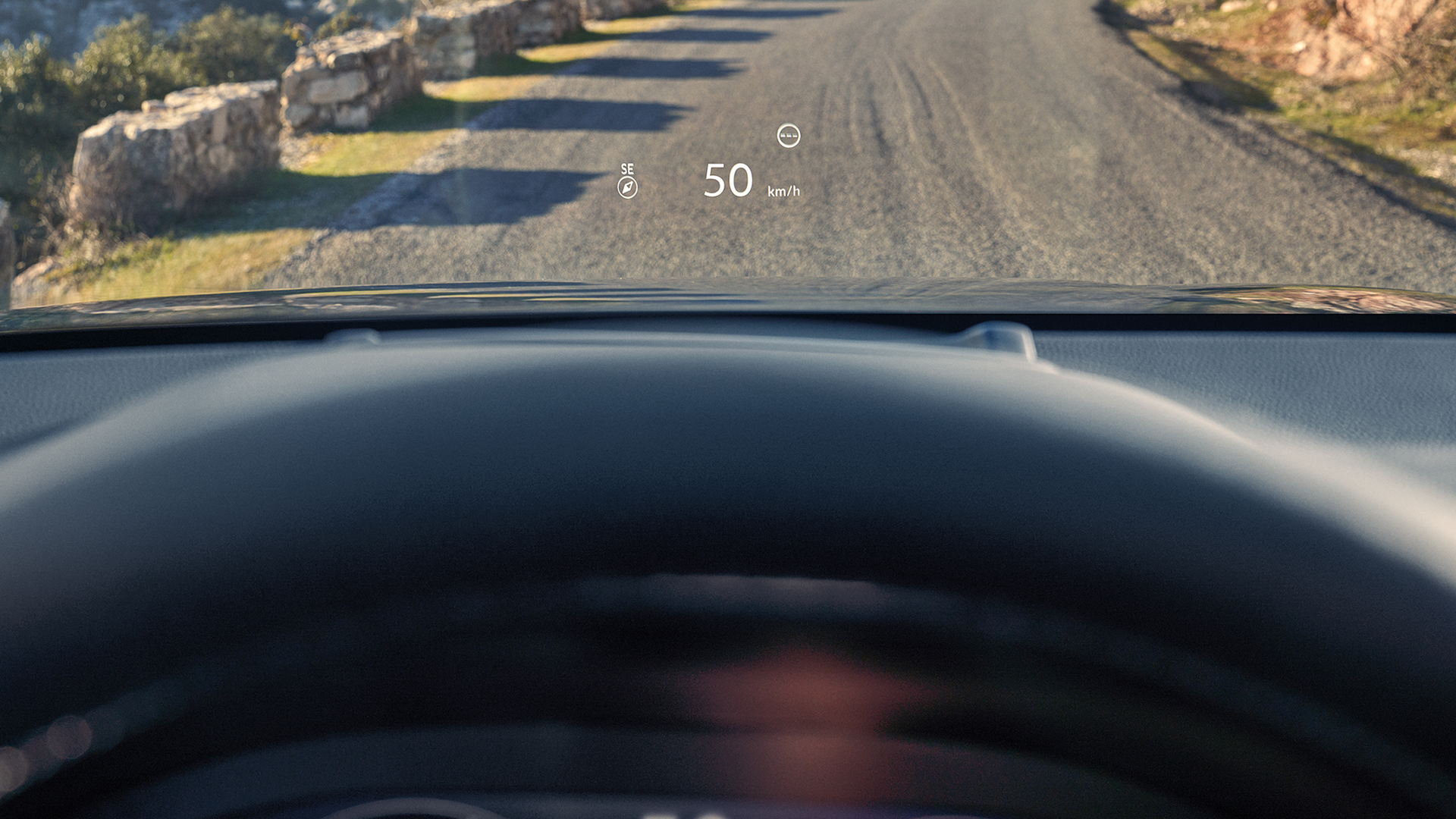 Vista en primera persona desde el asiento del conductor, mostrando un Head Up Display (HUD) que proyecta "50 km/h" y una brújula en el parabrisas sobre un camino de grava.