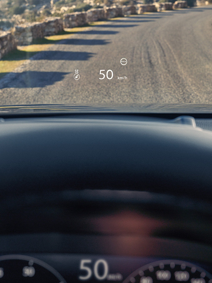 Vista en primera persona desde el asiento del conductor, mostrando un Head Up Display (HUD) que proyecta "50 km/h" y una brújula en el parabrisas sobre un camino de grava.