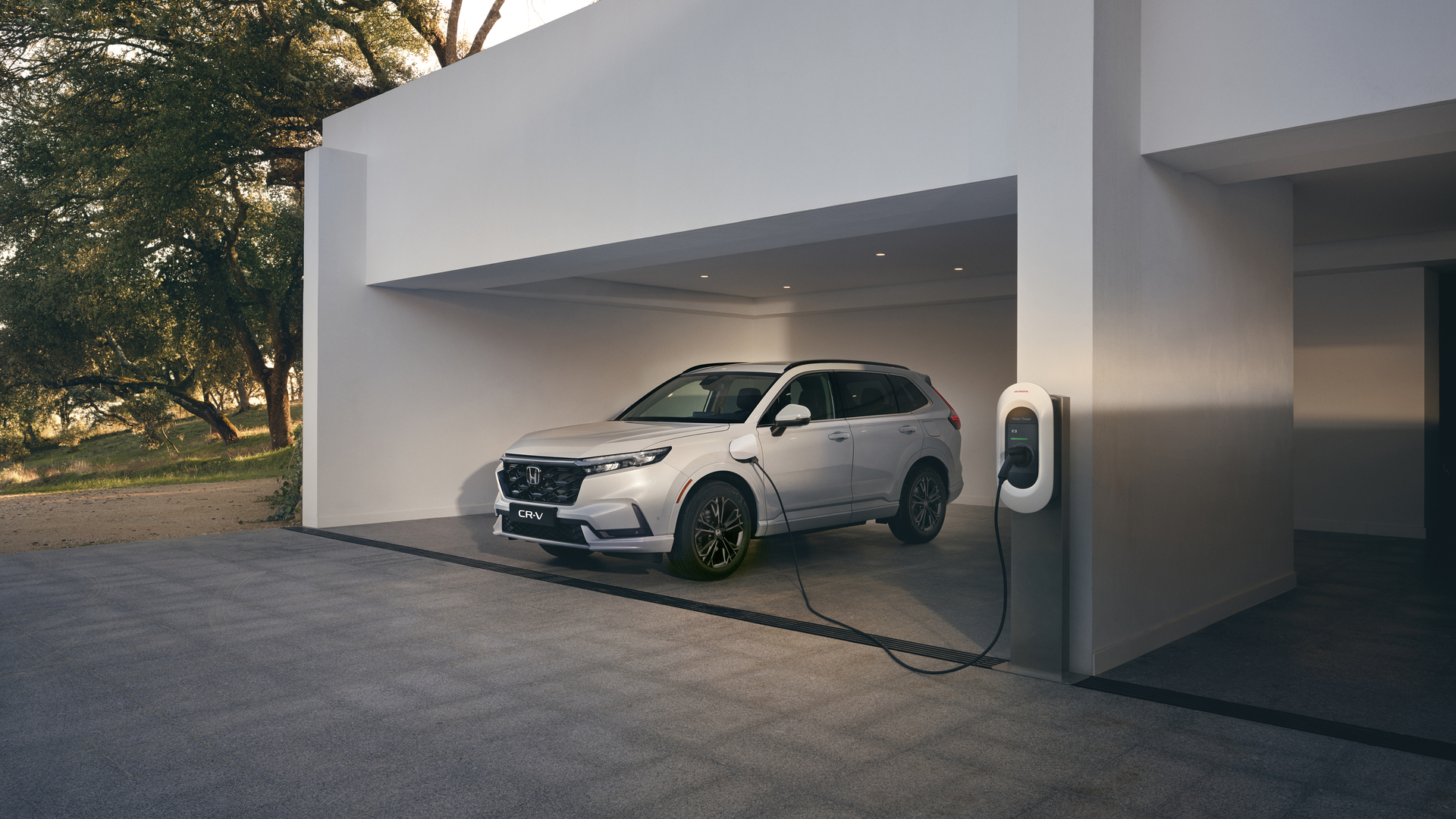 Un Honda CR-V Plug-in Hybrid blanco está aparcado en una cochera minimalista blanca, conectado a un cargador de vehículo eléctrico montado en la pared.