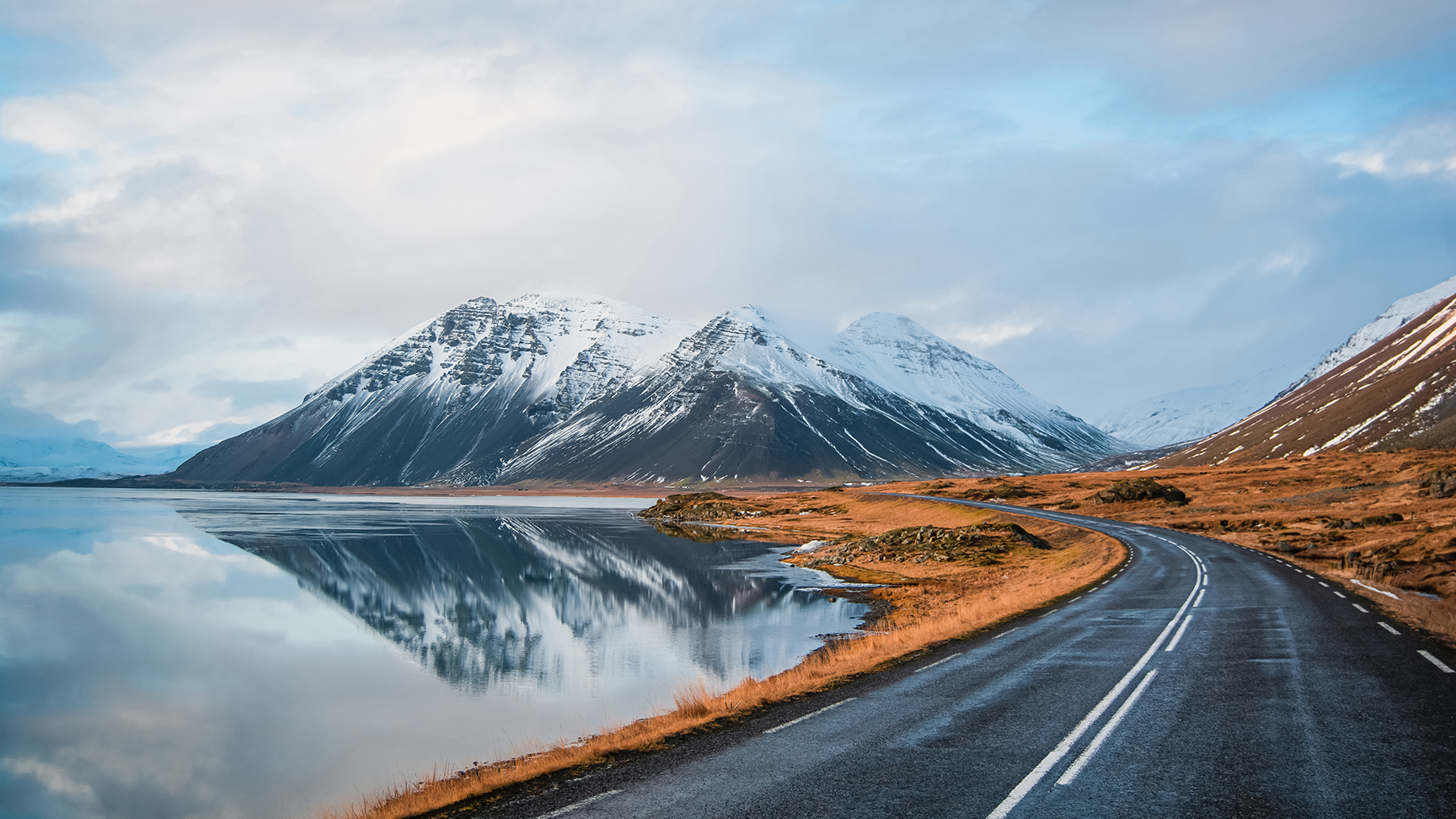 Foto panorámica de invierno de la carretera que conduce a lo largo de la costa del lago hasta las montañas volcánicas. Altos picos rocosos cubiertos de nieve que se reflejan en la superficie del agua. Punto de vista del conductor en la carretera circular de Islandia.