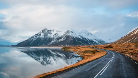 Foto panorámica de invierno de la carretera que conduce a lo largo de la costa del lago hasta las montañas volcánicas. Altos picos rocosos cubiertos de nieve que se reflejan en la superficie del agua. Punto de vista del conductor en la carretera circular de Islandia.