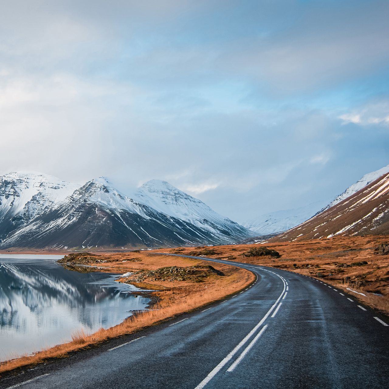 Foto panorámica de invierno de la carretera que conduce a lo largo de la costa del lago hasta las montañas volcánicas. Altos picos rocosos cubiertos de nieve que se reflejan en la superficie del agua. Punto de vista del conductor en la carretera circular de Islandia.