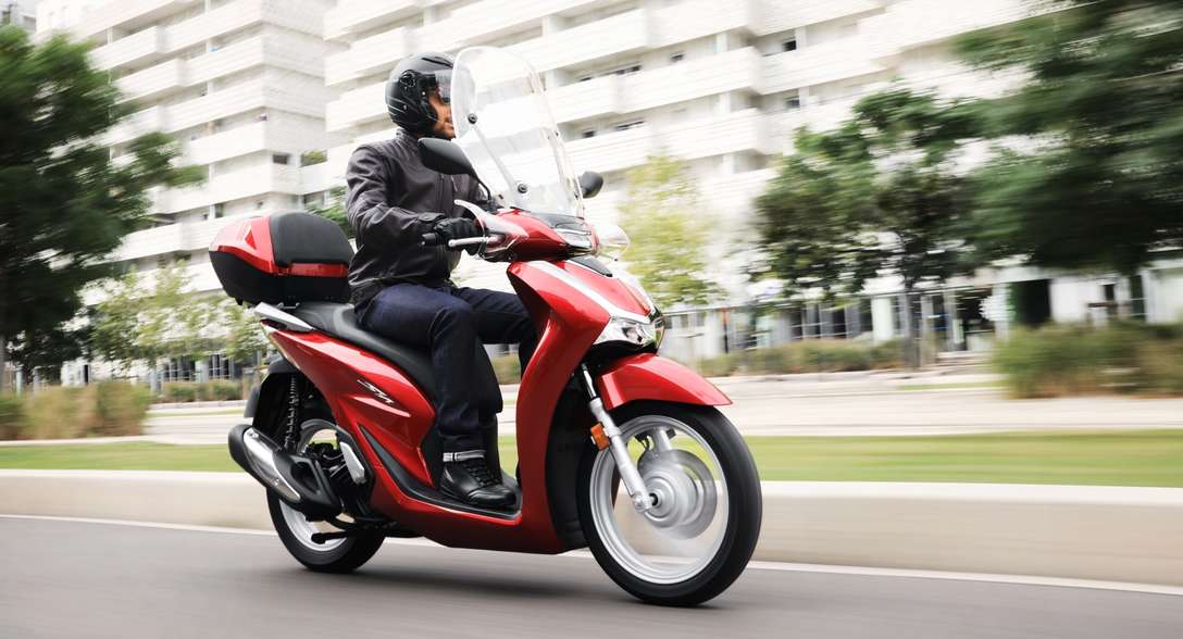 Honda-SH125i, vista de tres cuartos de la parte delantera derecha, con conductor, en la ciudad
