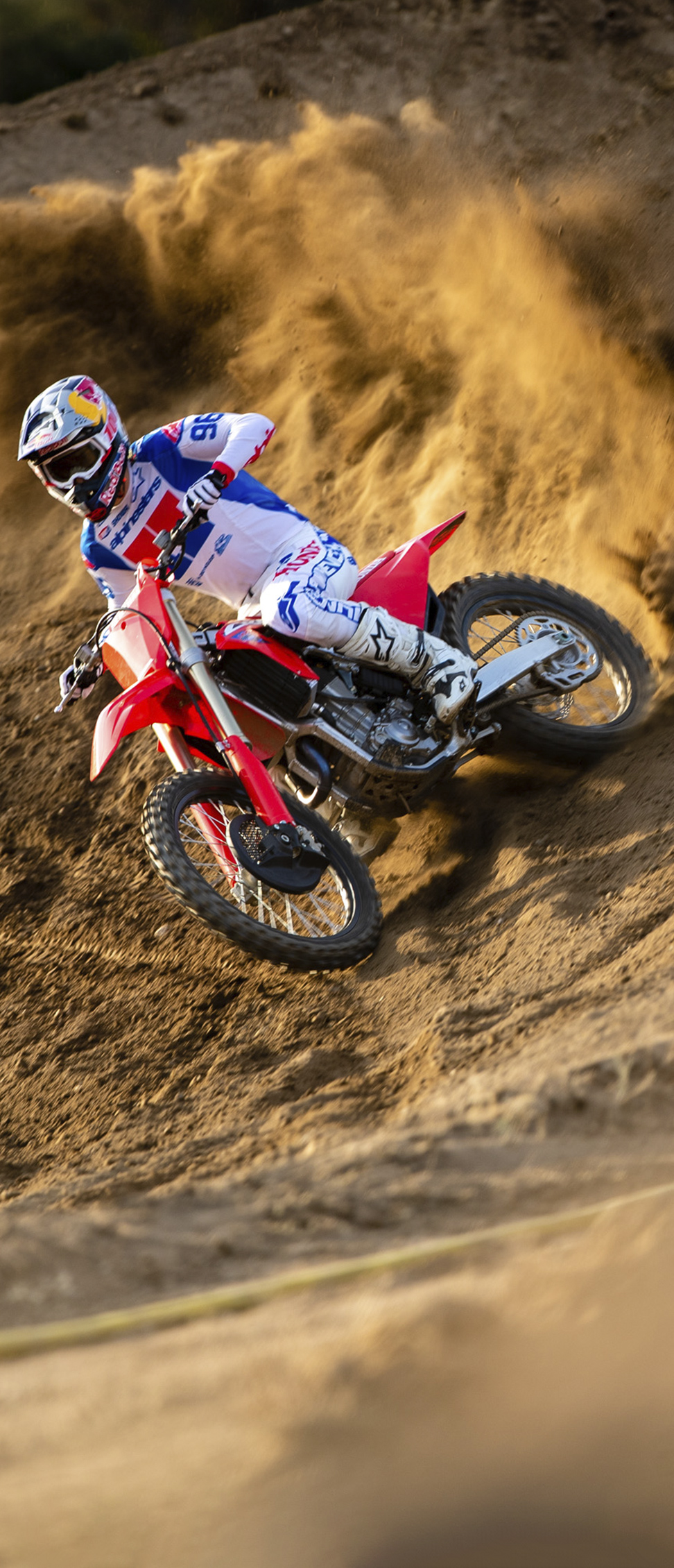 Un piloto de motocross, vestido con equipo blanco, azul y rojo, inclina una moto de cross roja en una curva arenosa pronunciada, levantando una gran ola de tierra mientras acelera en la vuelta.