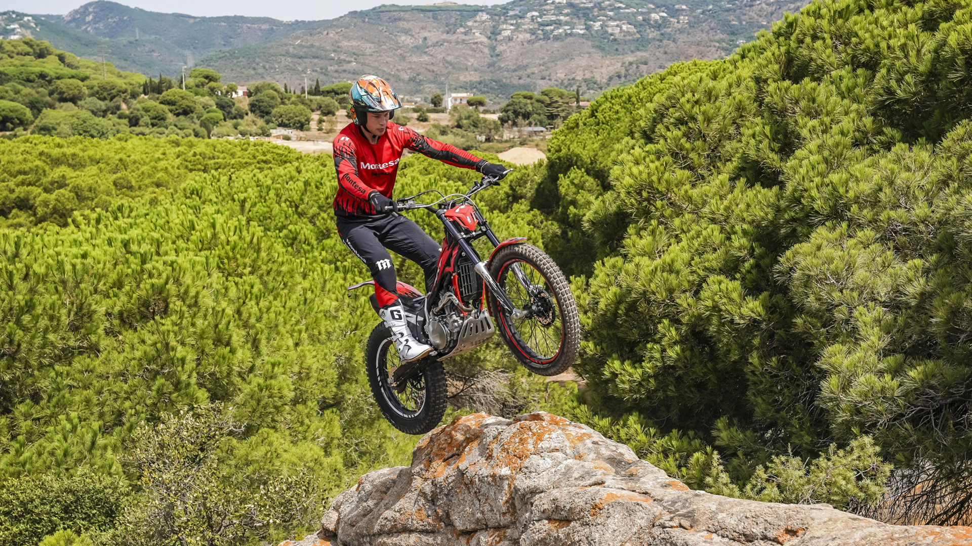 Conductor saltando con una Montesa Cota 4RT 260R roja desde una cornisa rocosa sobre un paisaje pintoresco de bosque de pinos.