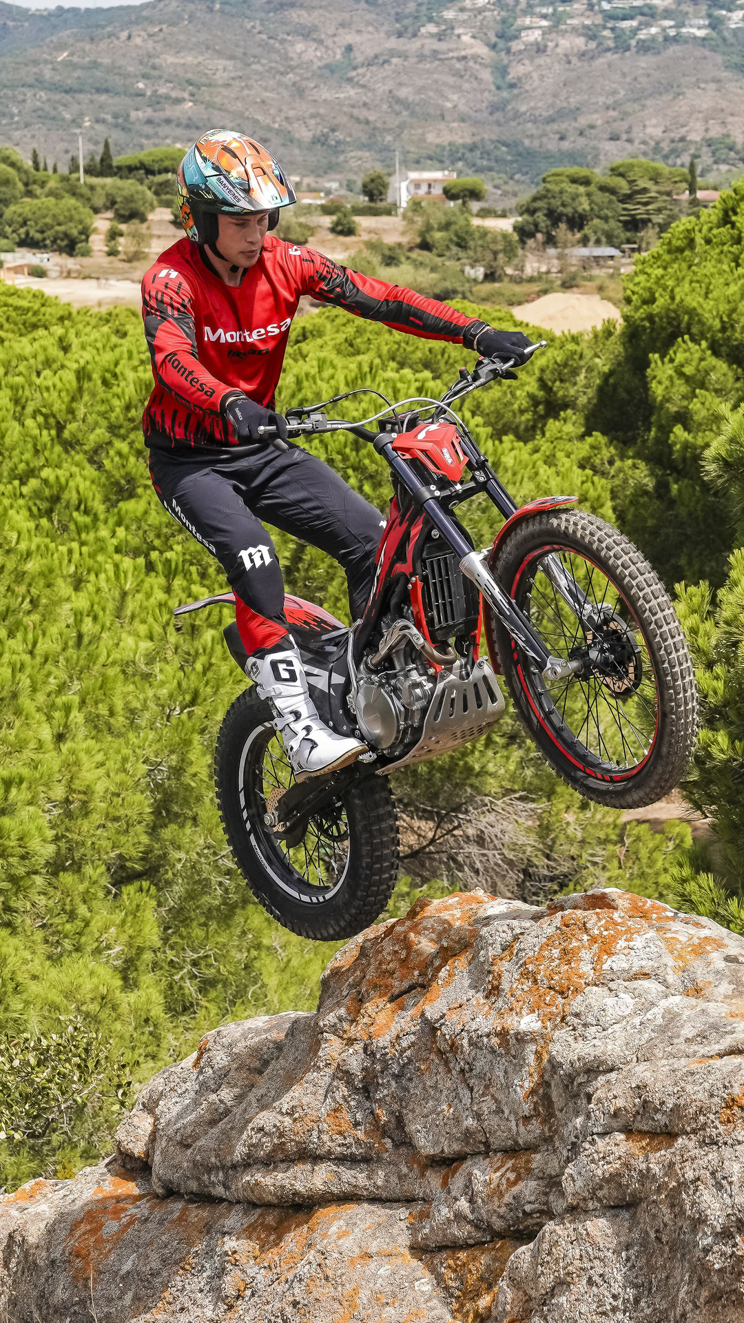 Conductor saltando con una Montesa Cota 4RT 260R roja desde una cornisa rocosa sobre un paisaje pintoresco de bosque de pinos.