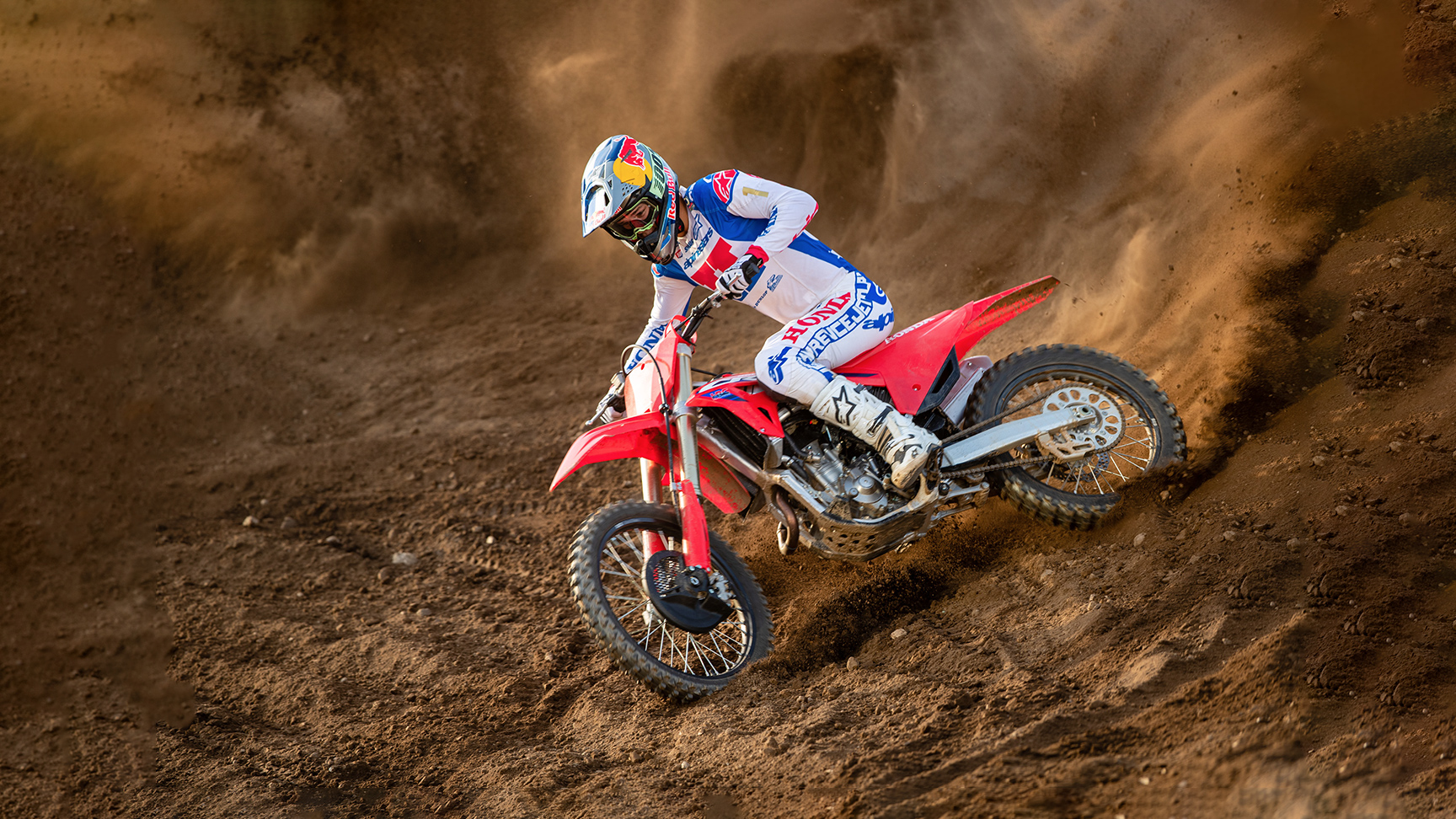 Un piloto de motocross, equipado con vestimenta de competición blanca, azul y roja, se inclina bruscamente en una curva sobre una moto de cross roja, recorriendo una pista arenosa y levantando una espectacular nube de tierra tras de sí.