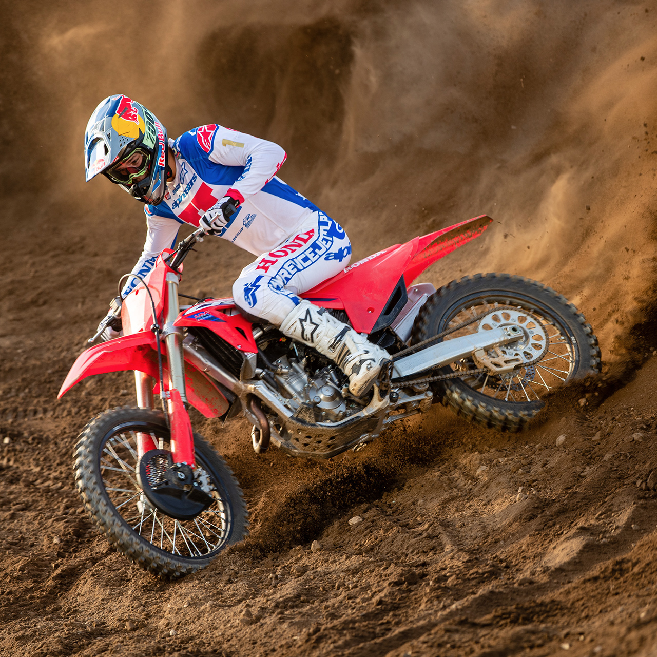 Un piloto de motocross, equipado con vestimenta de competición blanca, azul y roja, se inclina bruscamente en una curva sobre una moto de cross roja, recorriendo una pista arenosa y levantando una espectacular nube de tierra tras de sí.