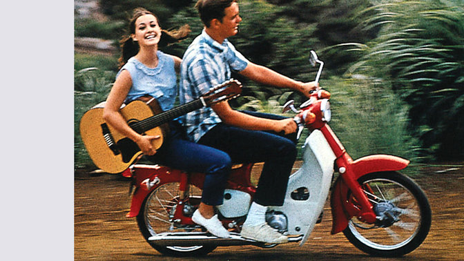 Honda Cub con conductor y un pasajero con una guitarra. Localización en una carretera polvorienta. Lado derecho.