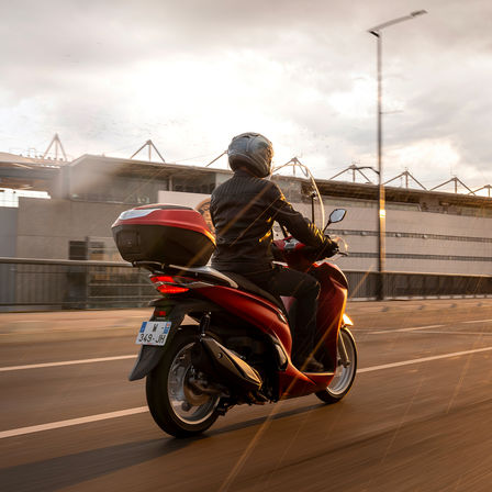 Honda SH350i, lateral derecho trasero de tres cuartos, con conductor, moto roja