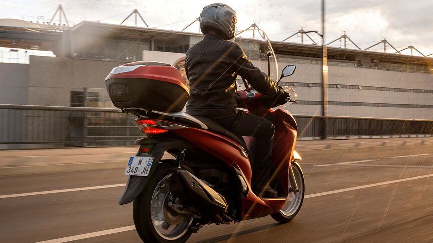 Honda SH350i, vista delantera derecha de tres cuartos, con conductor, en carretera, moto roja