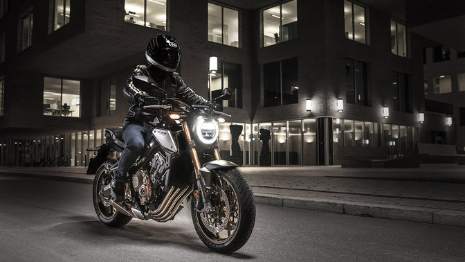 CB650R Neo Sports Café, lateral delantero derecho de tres cuartos con piloto, en un paisaje urbano de noche