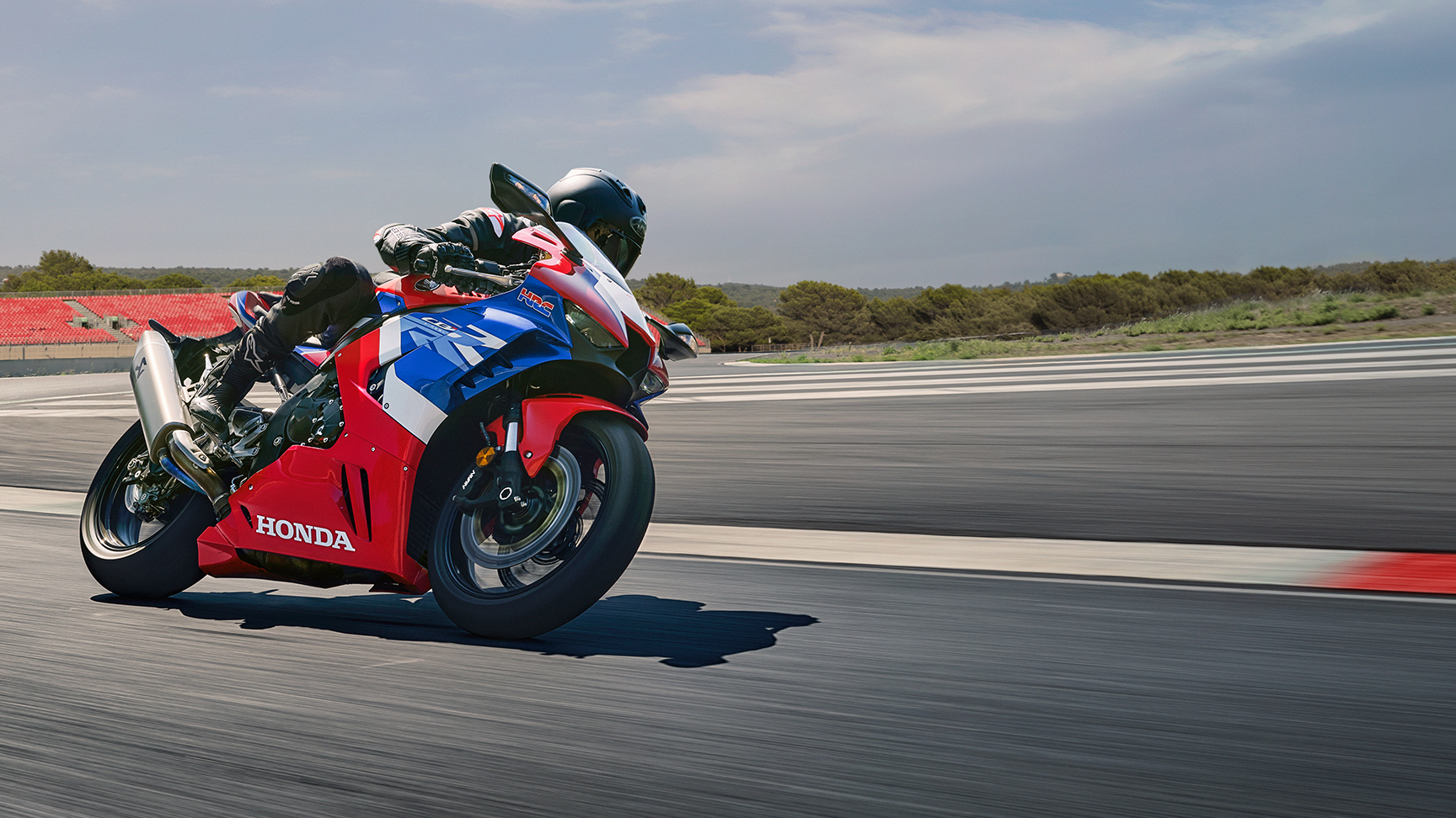 A rider in full black racing gear leans aggressively into a corner on a red, blue, and white Honda Fireblade sport motorcycle on a racetrack, with the asphalt blurred from speed and low greenery in the background.